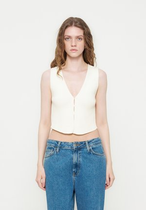 Femme aux longs cheveux ondulés portant un gilet court blanc sans manches et un jean bleu taille haute sur un fond uni.