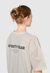Jasnoszara, oversize'owa koszulka z krótkim rękawem, z czarnym napisem na plecach "OFF DUTY GEAR" oraz dodatkowymi detalami druku.