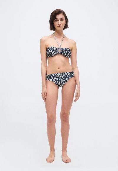 Γυναίκα στέκεται ξυπόλητη φορώντας μπικίνι σε σκούρο μπλε με λευκές οβάλ πουά και λουρί γύρω από το λαιμό, μπροστά σε απλό φόντο.