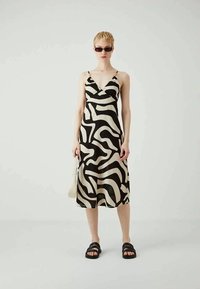 Vestido negro y beige con patrones ondulados, hecho de una tela suave. Presenta tirantes finos y un diseño de largo medio en forma de A. Se lleva con sandalias negras.