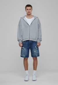 Pelēks zip-up hoody ar brīvu piegriezumu, kopā ar nodilušām zilo džinsu šortiem un baltām sporta kurpēm, attēlots uz neitrāla fona.