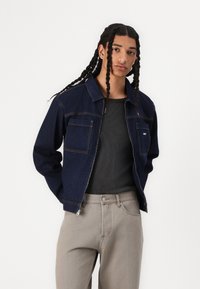 Denim jacket in dark blue with two front pockets, contrasted stitching, and a zip closure, worn over a grey ribbed shirt and light grey pants.