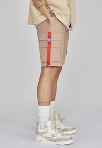 Pantaloni cargo beige con una cintura rossa e dettagli in fibbia argentata, due grandi tasche frontali e una texture morbida. Abbinati a sneakers bianche e beige.