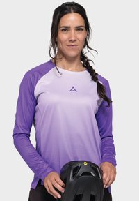 Lila Langarmshirt mit einem Farbverlauf-Design, das über eine atmungsaktive Textur verfügt und ein Logo in der Mitte hat. Schwarzer Helm, der in der linken Hand gehalten wird.