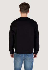 Hombre con cabello castaño rizado, vestido con una sudadera negra y jeans azules, de espaldas frente a un fondo gris claro liso.