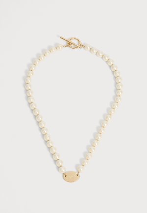 Collier de perles avec fermoir à bascule en or et pendentif ovale en or gravé de lettres sur fond blanc uni.