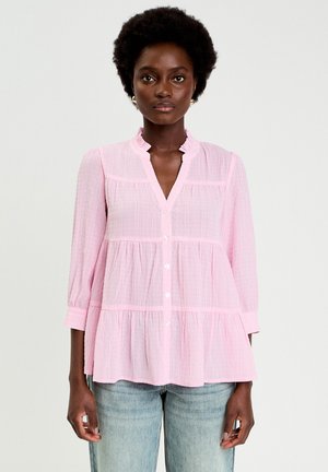 Blouse rose clair à rayures blanches verticales, manches longues, col à volants, boutonnage devant et ourlet évasé. Tissu doux et coupe décontractée.
