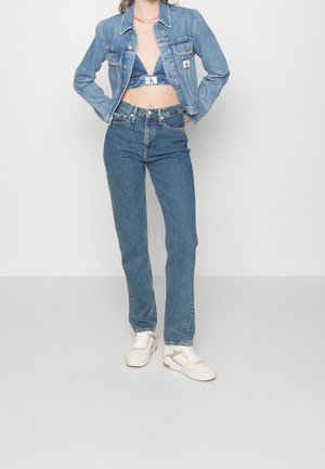 Persoon die een blauwe spijkerjasje draagt, bijpassend blauw spijkerjasje-bh-topje, hoge taille blauwe spijkerbroek en witte sneakers, met handen in de zij staand.