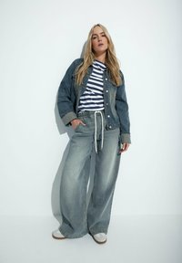 Giubbotto di jeans con bottoni, t-shirt a righe blu navy e bianche, jeans a gamba larga in denim chiaro con coulisse, e scarpe grigie con suole marroni.