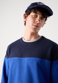 Navy and blue color-blocked sweatshirt with a textured finish, round neckline, and ribbed cuffs. Paired with a navy cap featuring white logo.