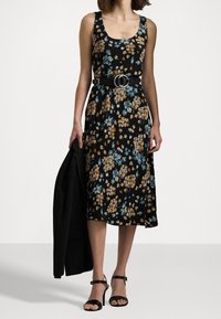 Kvinna i en ärmlös svart blommig midiklänning med ett svart bälte, som håller en svart kavaj och har på sig svarta högklackade sandaler.