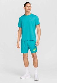 Camisa deportiva turquesa con un logo blanco, combinada con pantalones cortos a juego que presentan un texto en neón verde "RUN" y el swoosh de Nike, complementada con zapatillas blancas.