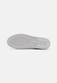 Suola di sneaker bianca con una superficie liscia, una texture sottile e bordi arrotondati. Presenta una piastra con logo incastonata vicino al tallone.