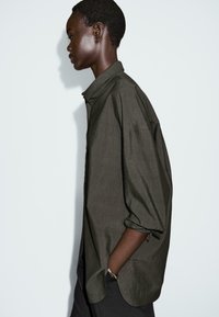 Camicia abbottonata oversize verde oliva con colletto a punta, maniche lunghe arrotolate ai polsini e orlo curvato. Tessuto testurizzato.