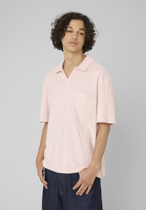 Jeune homme aux cheveux bouclés portant un polo rose clair à manches courtes et un jean bleu foncé sur un fond gris uni.