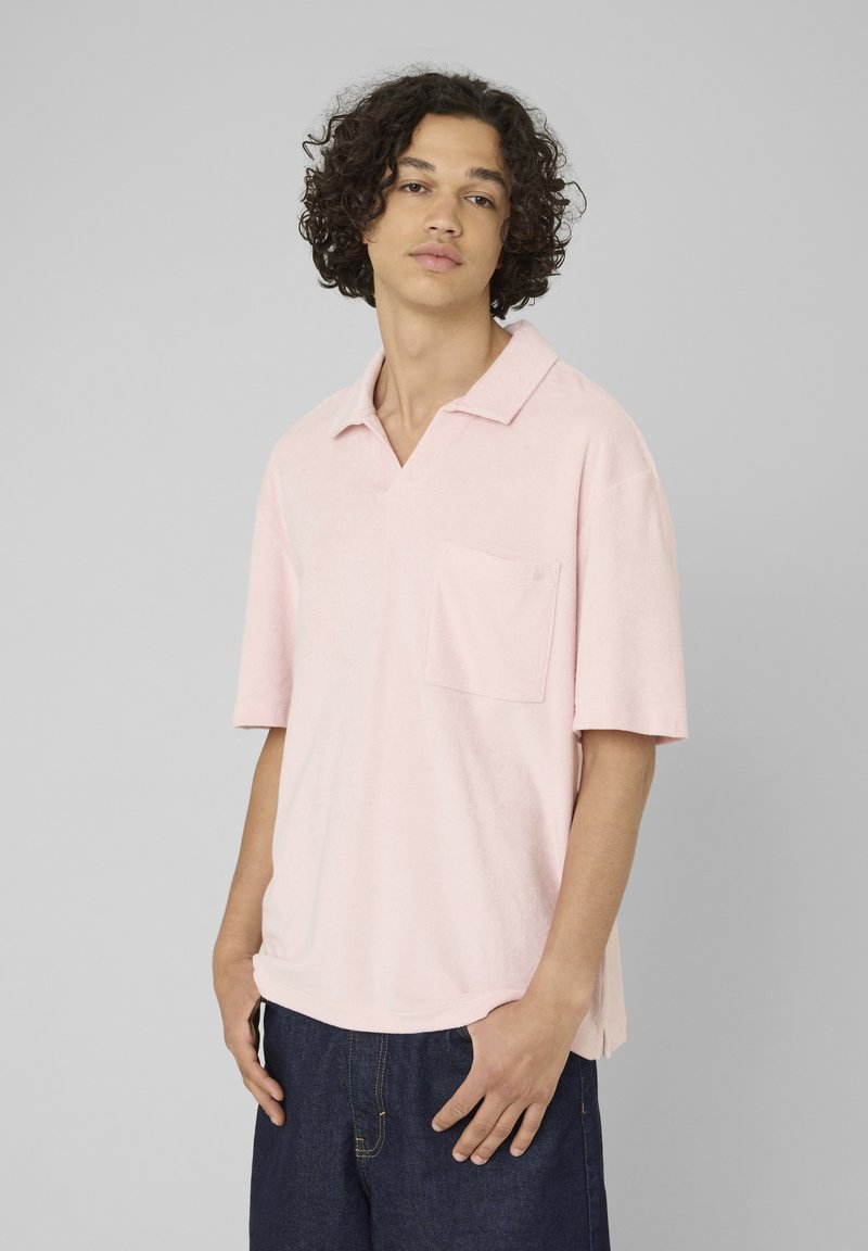 Jeune homme aux cheveux bouclés portant un polo rose clair à manches courtes et un jean bleu foncé sur un fond gris uni.