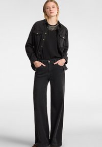 Camisa de mezclilla negra con cierre de botones en la parte delantera y dos bolsillos en el pecho, combinada con jeans negros de pierna ancha. Textura suave con diseño minimalista.