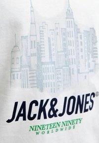 Witte stof met een lichtblauwe schets van een stadsgezicht boven vetgedrukte zwarte tekst "JACK & JONES" en kleinere groene tekst "NINETEEN NINETY WORLDWIDE".