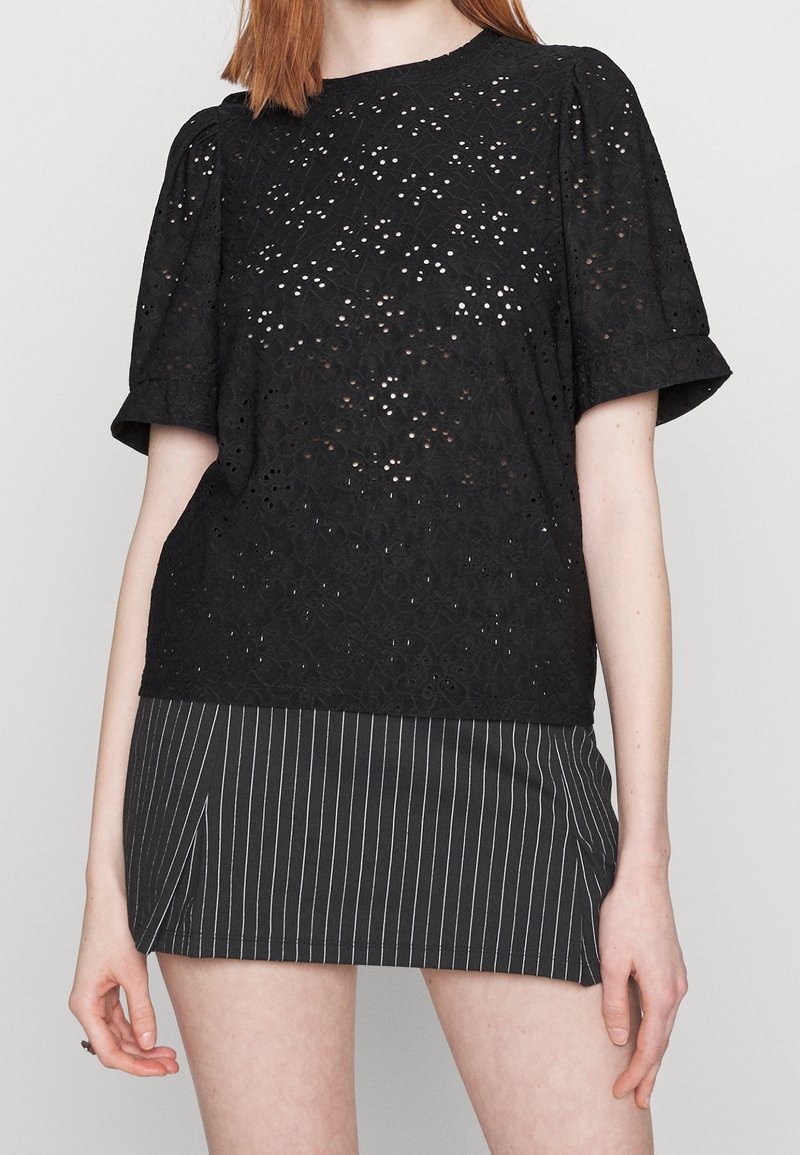 Haut en dentelle noire avec de courtes manches bouffantes, présentant des motifs découpés complexes. Assorti à une jupe noire à rayures fines. Design minimaliste.