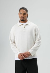 Long-sleeve cream pullover with a textured fabric, collar, and three-button placket. Worn over a white t-shirt, paired with black pants.