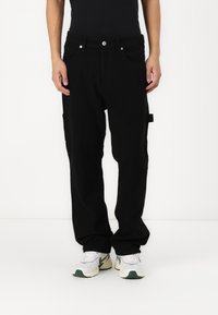 Pantaloni neri larghi in cotone, con un design a gamba dritta, tasche frontali e accessori metallici a vista. Indossati con sneakers bianche e verdi.