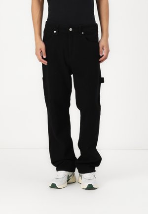 Black baggy pants made of cotton, featuring a straight leg design, front pockets, and visible metal hardware. Worn with white and green sneakers.