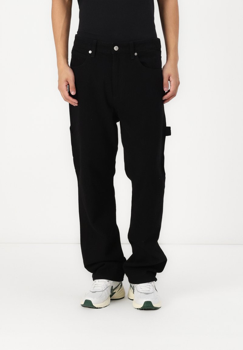Pantaloni neri larghi in cotone, con un design a gamba dritta, tasche frontali e accessori metallici a vista. Indossati con sneakers bianche e verdi.