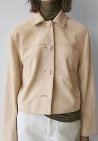 Beige ruskindjakke med et cropped design, der har en krave og fire store knapper. Underneden skimtes en mørkegrøn højhalset trøje.