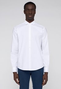 Witte knop-overhemd gemaakt van gladde stof, met een puntige kraag en lange mouwen. Model draagt het met donkerblauwe spijkerbroek.