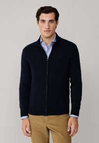 Mornarska zip-up jopica iz teksturiranega materiala, z visokim ovratnikom in gubanimi manšetami, preoblečena čez svetlo modro prugasto srajco.