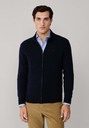 Suéter azul marino con cremallera, hecho de tela texturizada, presenta un cuello alto y puños acanalados, superpuesto a una camisa a rayas azul claro.