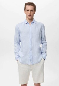 Camisa de lino de manga larga azul claro con cuello abotonado, que presenta un ajuste ligeramente relajado y una tapeta frontal limpia.