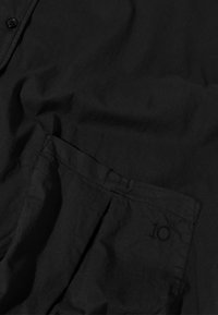 Chemise noire à boutons en tissu doux, avec une poche poitrine ornée d'un discret chiffre brodé "10" et une coupe décontractée.