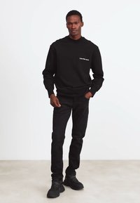 Felpa nera con scollo arrotondato, caratterizzata da un marchio bianco. Abbinata a jeans neri e calzature scure. Colore uniforme, design casual.