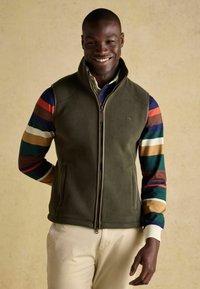 Kamizelka z polaru w kolorze oliwkowym, z bocznymi kieszeniami. Pod spodem znajduje się koszula w paski w wielokolorowym wzorze, z niebieskimi, czerwonymi, zielonymi i brązowymi paskami.