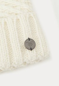 Knitted cream beanie hat with thick ribbing, featuring a round metallic logo tag sewn onto the side with black stitching.