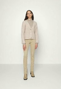 Mulher de pé contra um fundo liso, vestindo um cardigan bege, calças combinando e botins de salto médio castanho claro.