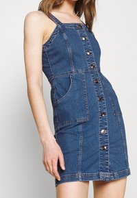 Kvinna i en ärmlös blå denimklänning med knappar framtill och stora sidofickor, stående mot en enkel vit bakgrund.