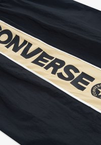 Tissu noir avec une grande bande beige portant le mot « CONVERSE » en lettres noires majuscules ; comprend un logo rond avec une étoile.