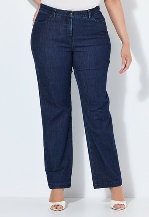 Plus-Size-Person, die dunkelblaue Jeans mit geradem Bein und weiße, offene Sandalen mit Absätzen vor einem einfarbigen Hintergrund trägt.