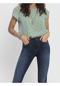 Femme portant un t-shirt vert à manches courtes avec des taches blanches, un jean bleu foncé, un collier en chaîne avec une croix, un bracelet et des bagues, mains dans les poches.