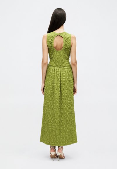 Γυναίκα με μακριά σκοτεινά μαλλιά που φοράει ένα πράσινο μάξι φόρεμα χωρίς μανίκια με υφή και άνοιγμα στην πλάτη, στέκεται με ψηλοτάκουνα σε ένα απλό λευκό φόντο.