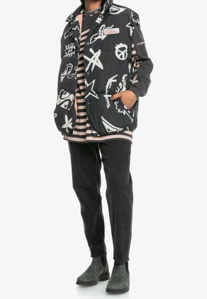 Personne portant un gilet matelassé noir avec des motifs abstraits blancs sur une chemise à manches longues rayée rose et noir, un jean noir et des bottines foncées.