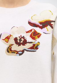 Gros plan sur un tissu blanc avec des fleurs abstraites brodées dans des nuances de rouge, beige, marron, jaune et bleu clair sur un vêtement.