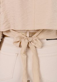 Beige top met een gestructureerde uitstraling, vastgebonden in de taille met een bijpassende stoffen sjaal over lichtgekleurde, aansluitende broeken. Zichtbaar detail met gouden hardware.