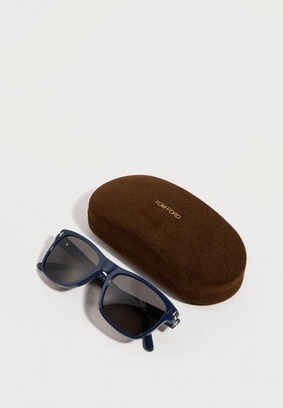 Blue square-frame sunglasses with dark lenses beside a brown Tom Ford branded hard case on a white background.