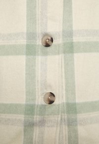 Kostkovaná košile se zelenými a šedými vzory na světle béžovém pozadí, s dvěma kulatými vzory zdobenými knoflíky a měkkou, strukturovanou látkou.