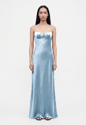 Femme portant une longue robe en satin bleu clair avec des fines bretelles et un détail structuré au niveau du buste, debout devant un fond blanc uni.