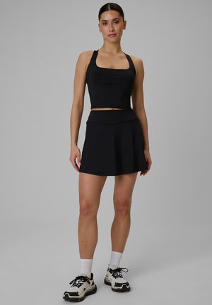 Kobieta ubrana w czarny bezrękawnik typu crop top, czarną spódnicę, białe skarpetki i białe trampki, stojąca na tle szarego tła.