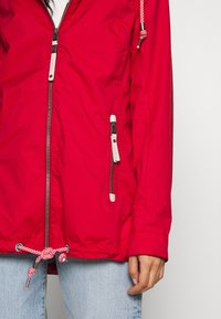 Červená voděodolná bunda se zipovým uzávěrem, šňůrkou na stažení v dolní části, bočními kapsami s kontrastními zipy a lehkým designem.